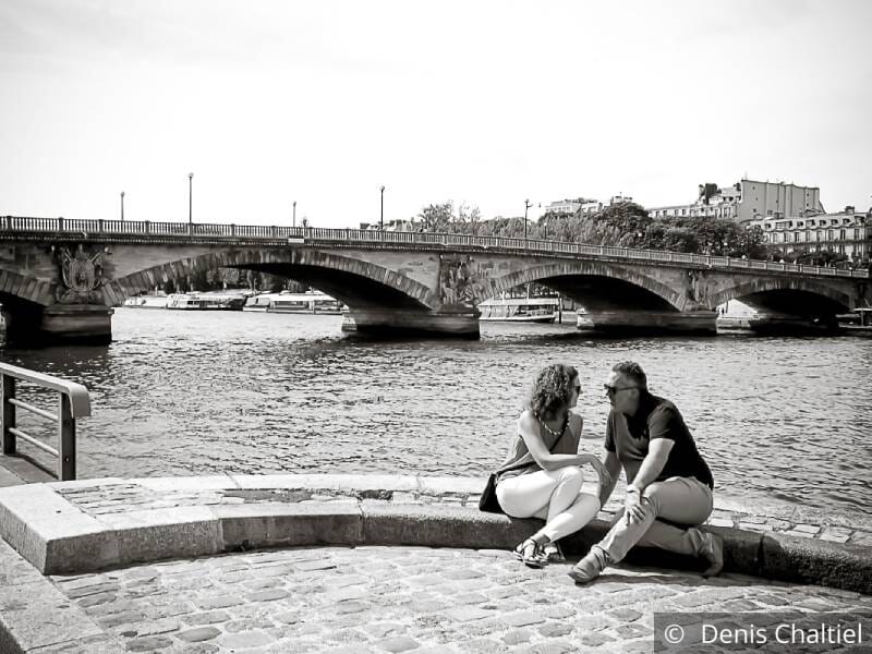 Un couple est assis au bord de l'eau et se regarde amoureusement. Photo en noir et blanc avec un pont en arrière plan.
